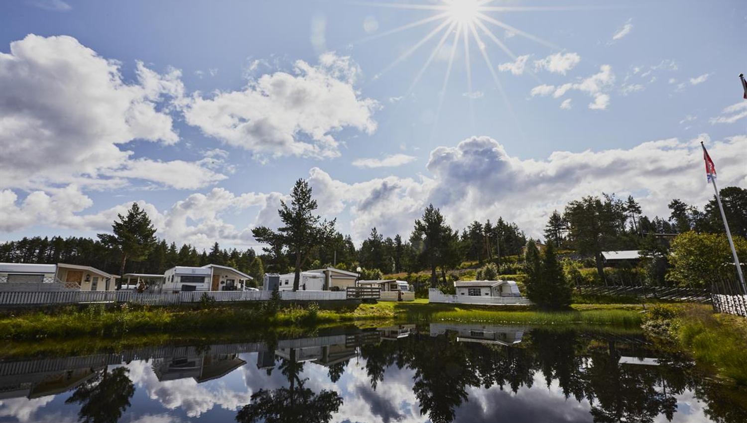 Campingplassen i strålende sol med noen få skyer på himmelen. Viser blikkstille vann i forkant av campingplassen.