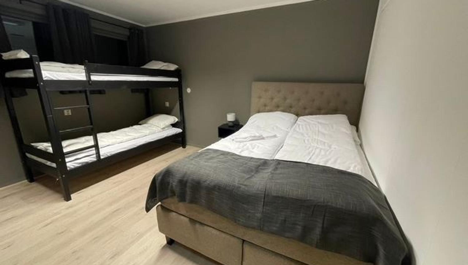 Compact bedroom with a double bed and dark throw, plus a black bunk bed, gray walls, and wood floor.