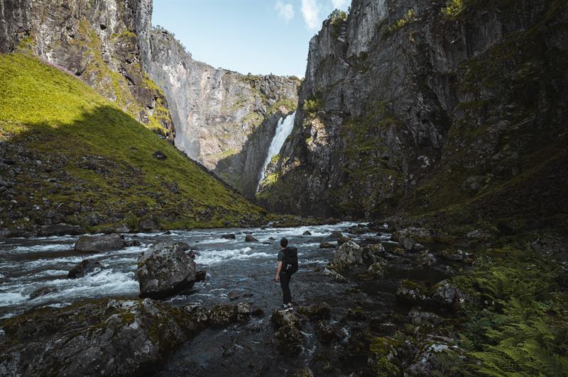 A man standing on the riverbank looking towards the waterfall.
