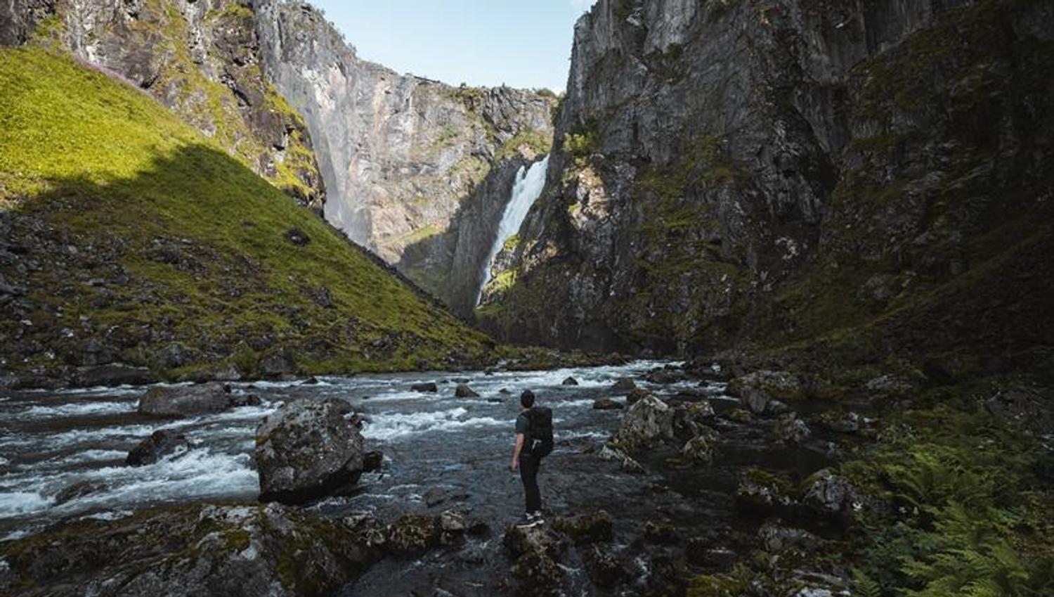 A man standing on the riverbank looking towards the waterfall.