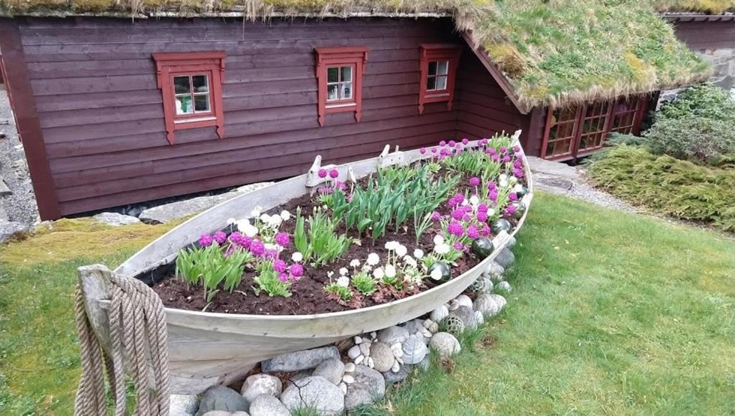 Orheimstunet in Nordfjord - A wooden boat used as a flower bed sits on rocks in a grassy yard beside a red cottage with a turf roof..