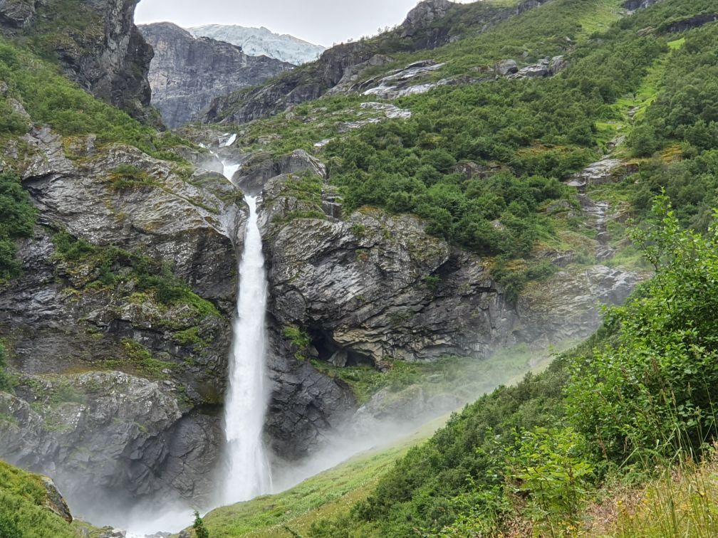 Tall waterfall plunges down dark cliffs into mist, surrounded by green shrubs and steep mountain slopes.