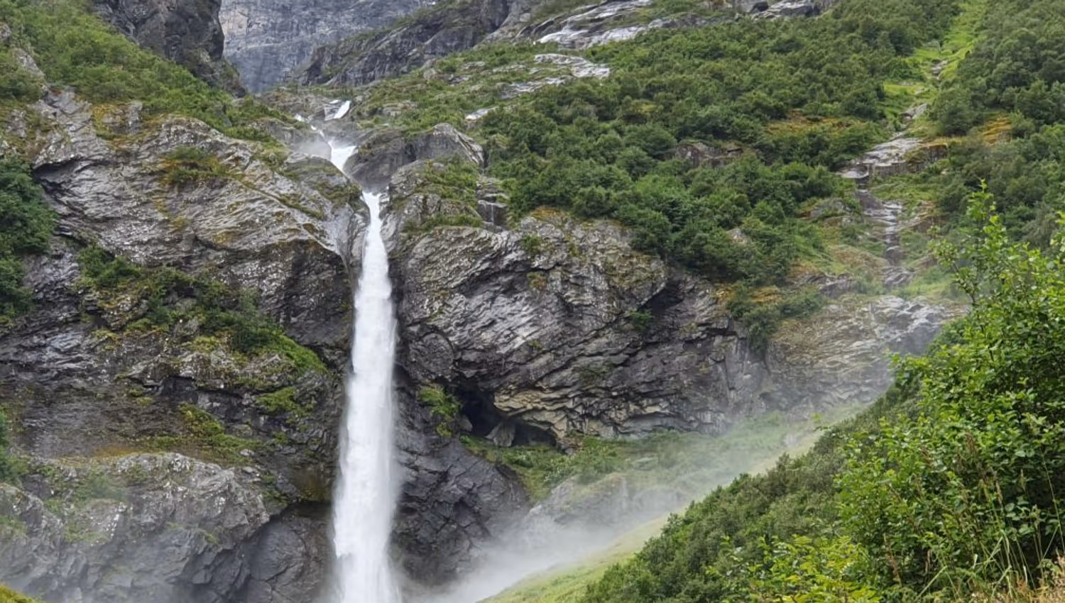 Tall waterfall plunges down dark cliffs into mist, surrounded by green shrubs and steep mountain slopes.