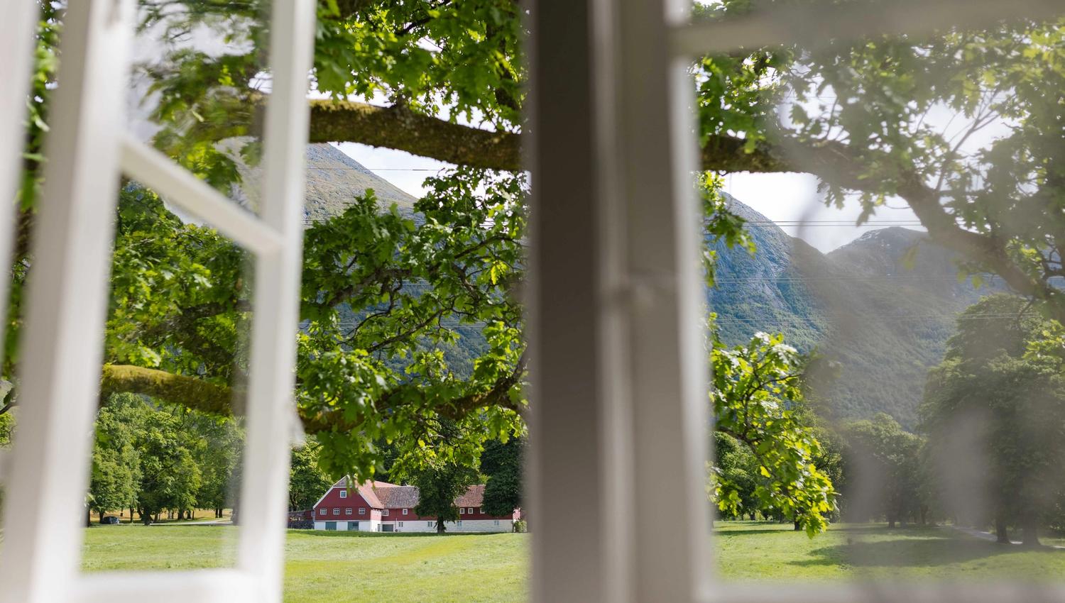 Utsikt frå vindauget i Fruehuset mot den historiske Avlsgården på Baroniet Rosendal, omgitt av grøne marker og majestetiske fjell.