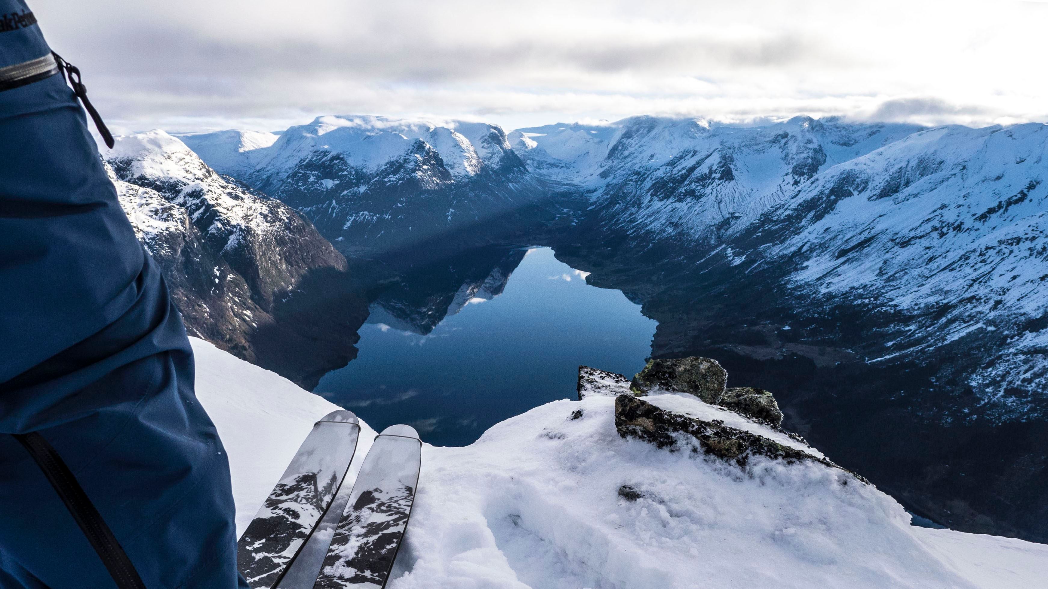 Skier’s legs and ski tips on a snowy ridge above a calm fjord, with rugged snow-covered mountains under clouds..