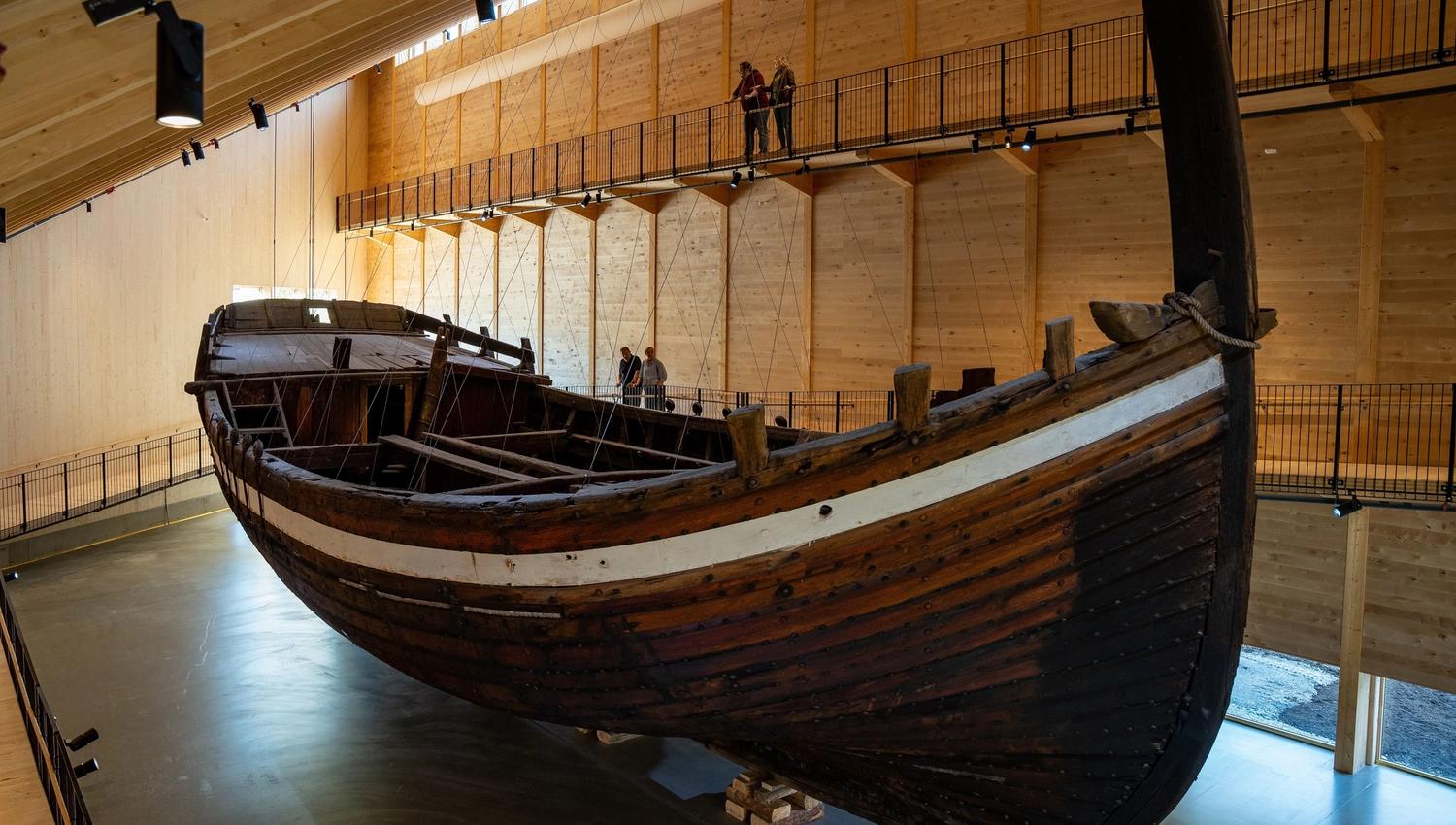 Large wooden boat, Holvikejekta, displayed indoors in a timber museum hall, with elevated walkways and a few visitors above..