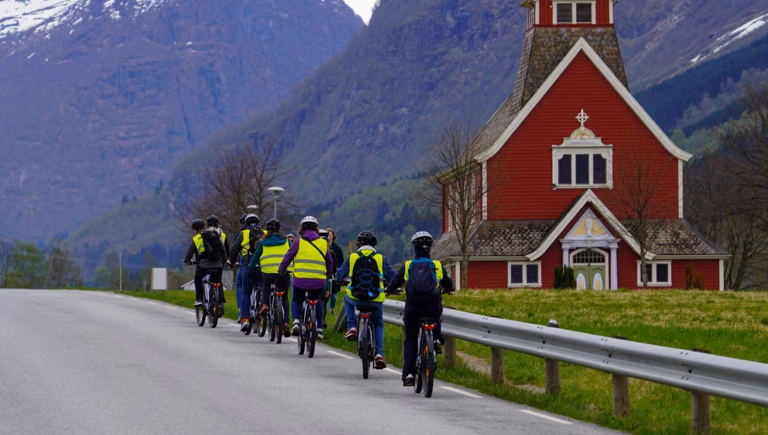 Group of cyclists on a rural road near a red church, with snowcapped mountains and cloudy sky behind.
