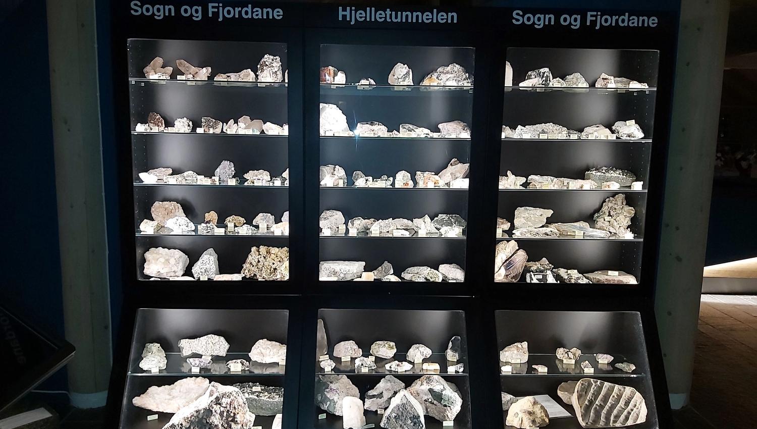 Svart utstillingsskap med opplyste hyller fulle av steinar og mineral, med skiltet «Sogn og Fjordane» og «Hjelletunnelen» øvst..
