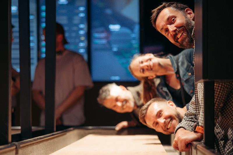 Smiling friends leaning over a shuffleboard table, watching the puck in a social gaming environment.