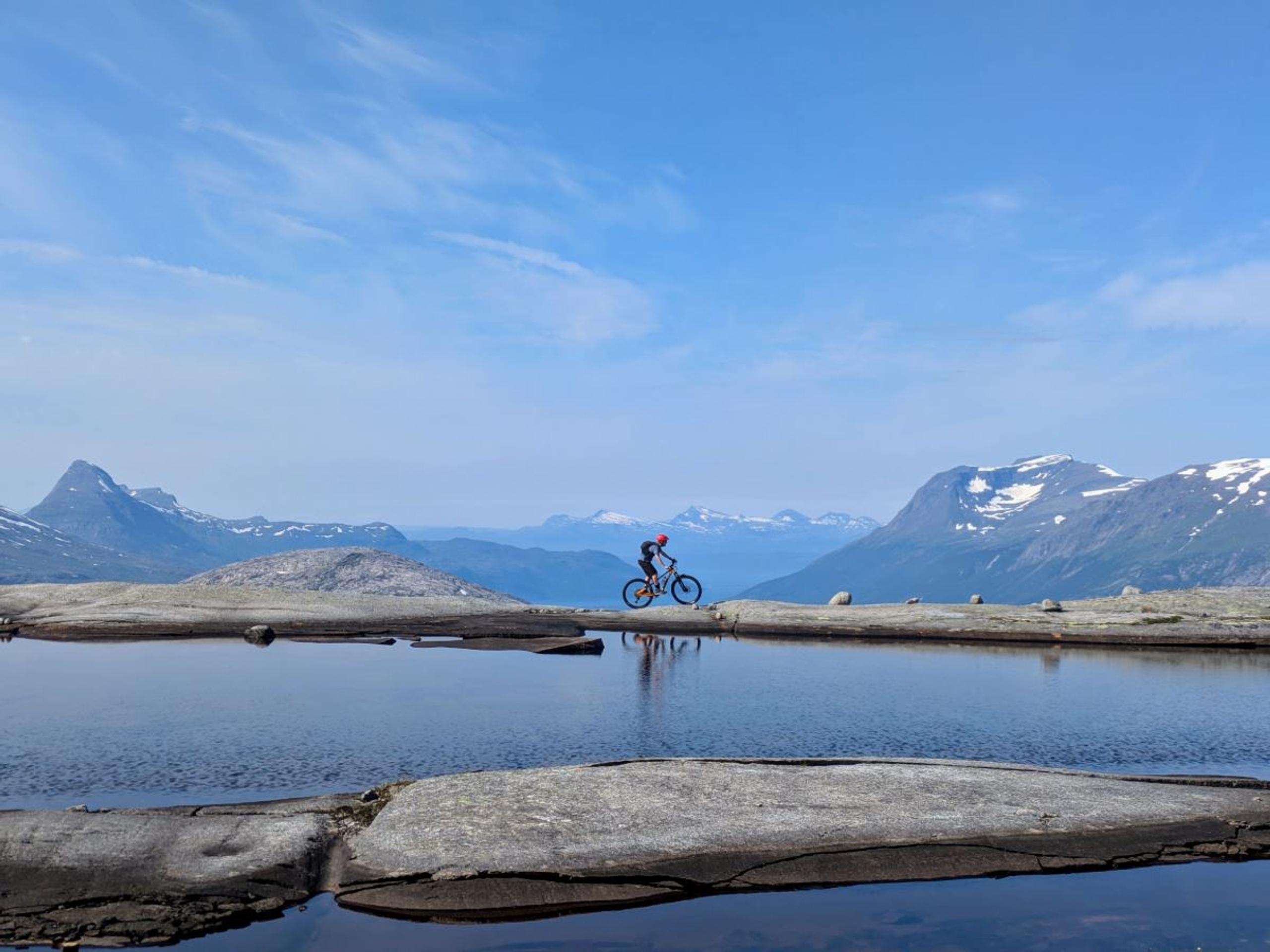 A lone cyclist rides along a rocky ridge above a calm lake, with distant snow-capped mountains under a blue sky.