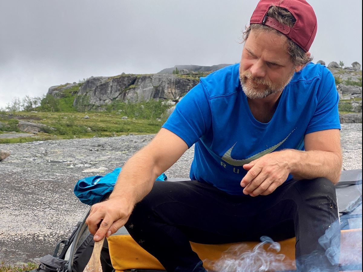 A person in a blue t-shirt and red cap sits outside by smoke from a small fire on a bedrock under a grey sky.
