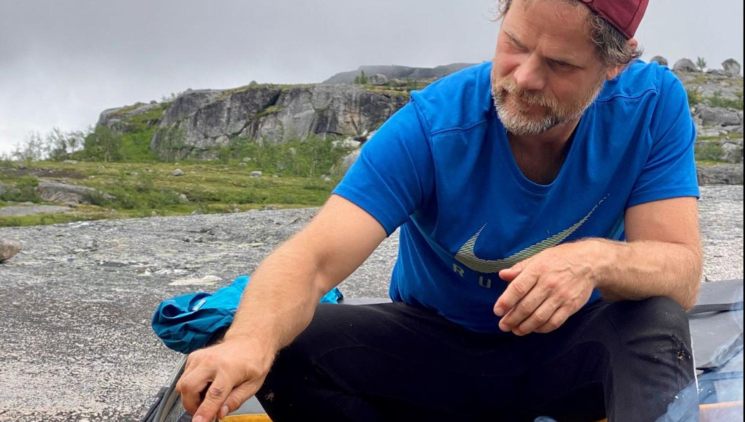 A person in a blue t-shirt and red cap sits outside by smoke from a small fire on a bedrock under a grey sky.