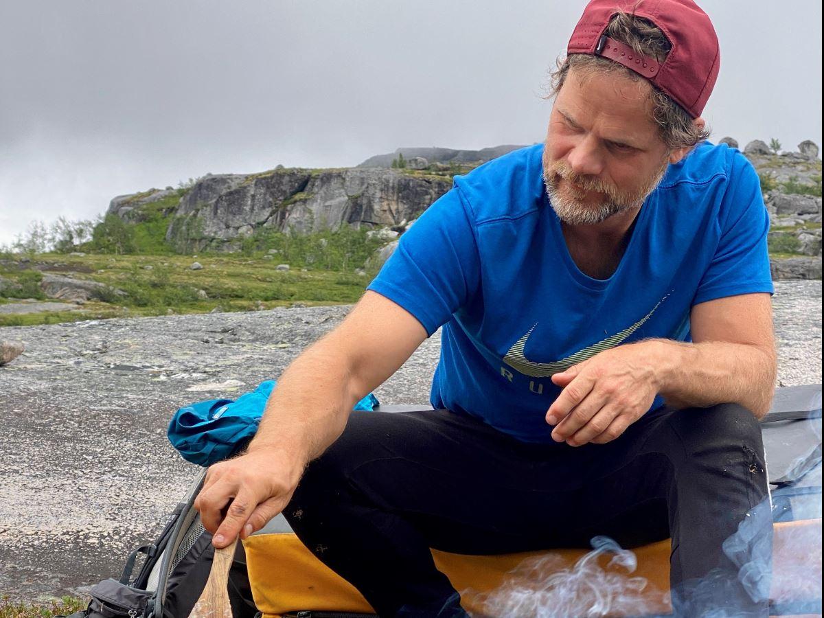 A person in a blue t-shirt and red cap sits outside by smoke from a small fire on a bedrock under a grey sky.