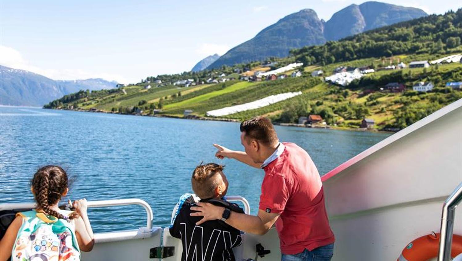 Far peiker mot frukthagar i fjordliene for barna sine på dekket under fjordcruise på Hardangerfjorden.