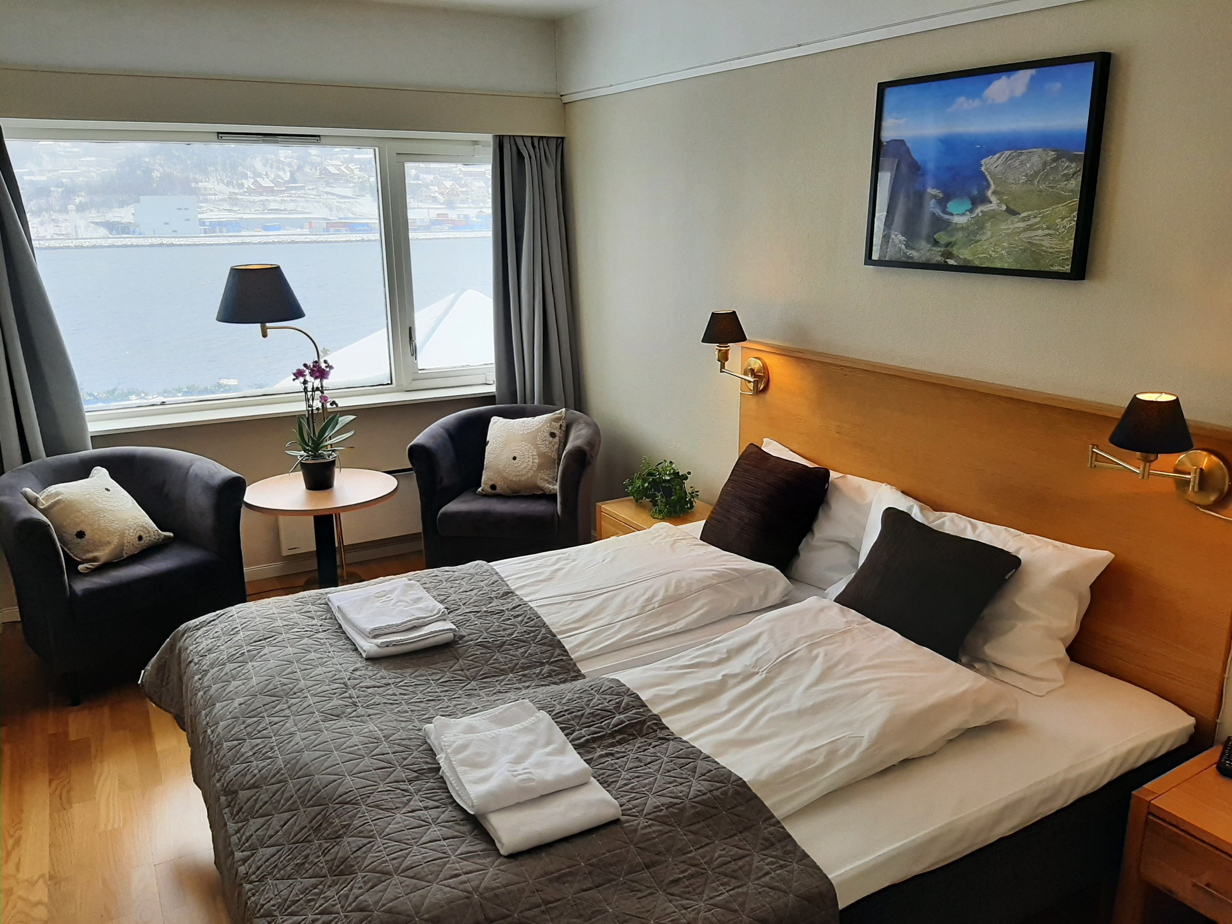 Eit lyst hotellrom på Bremanger Fjordhotell med dobbeltseng, to lenestolar ved vindauget, bord med plante og utsikt mot vatn..