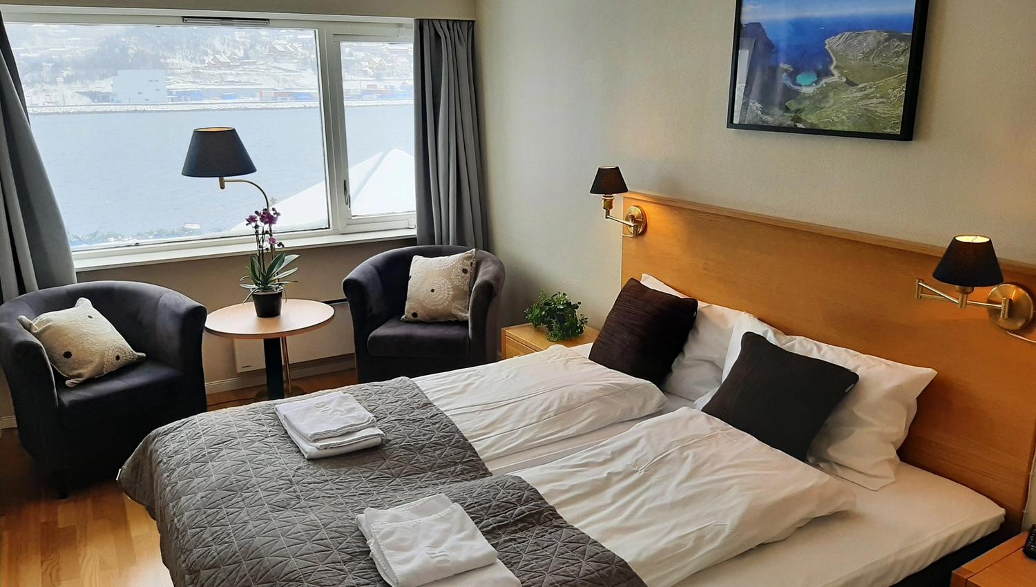 Eit lyst hotellrom på Bremanger Fjordhotell med dobbeltseng, to lenestolar ved vindauget, bord med plante og utsikt mot vatn..