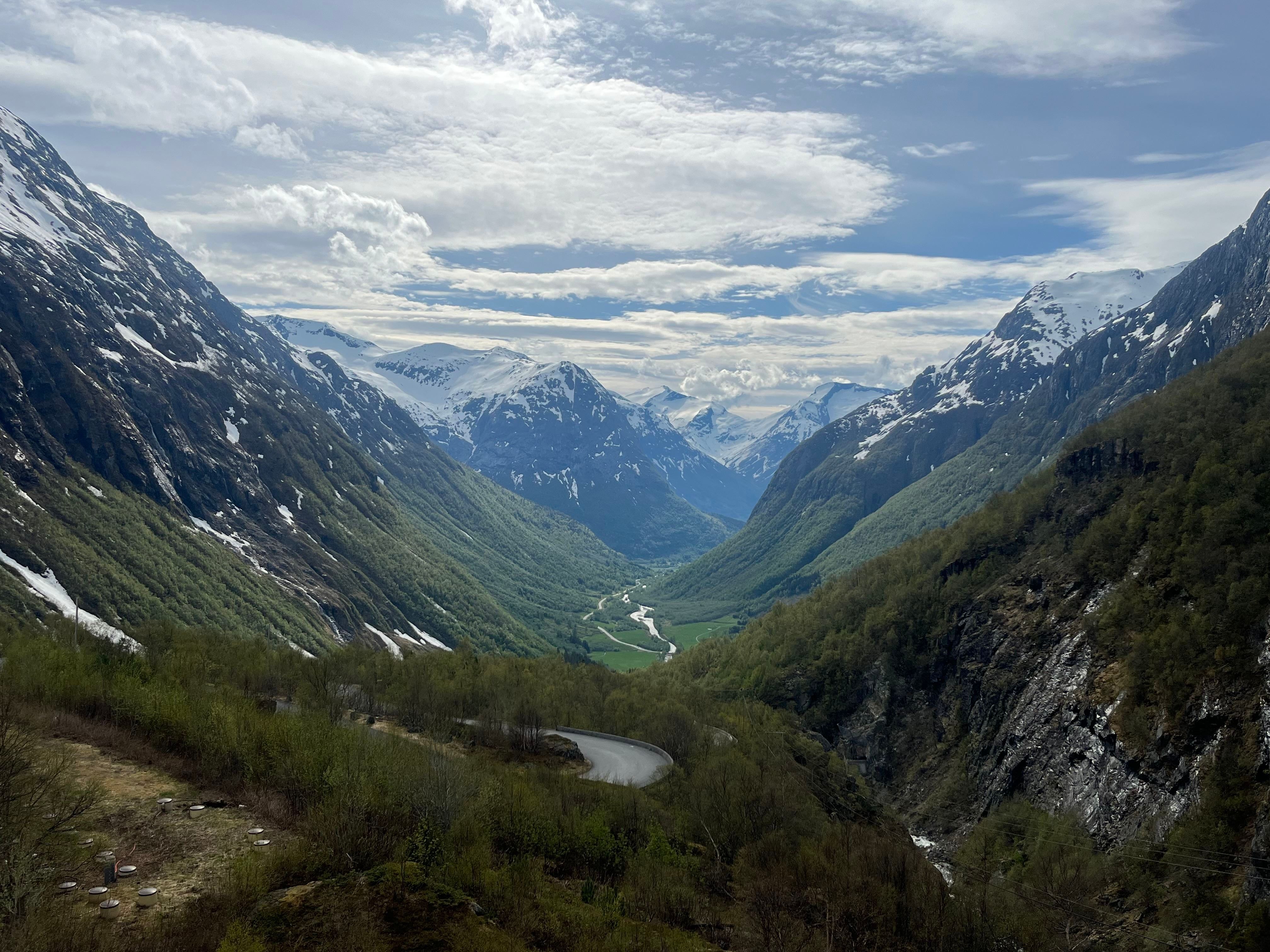 Wide view of a green mountain valley with snow-capped peaks, a winding road, and a river under cloudy sky..