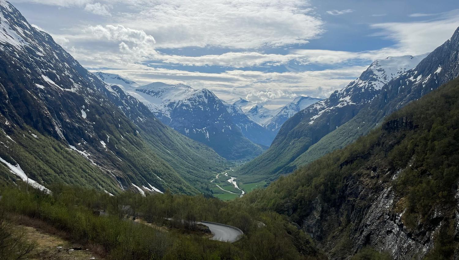 Wide view of a green mountain valley with snow-capped peaks, a winding road, and a river under cloudy sky..