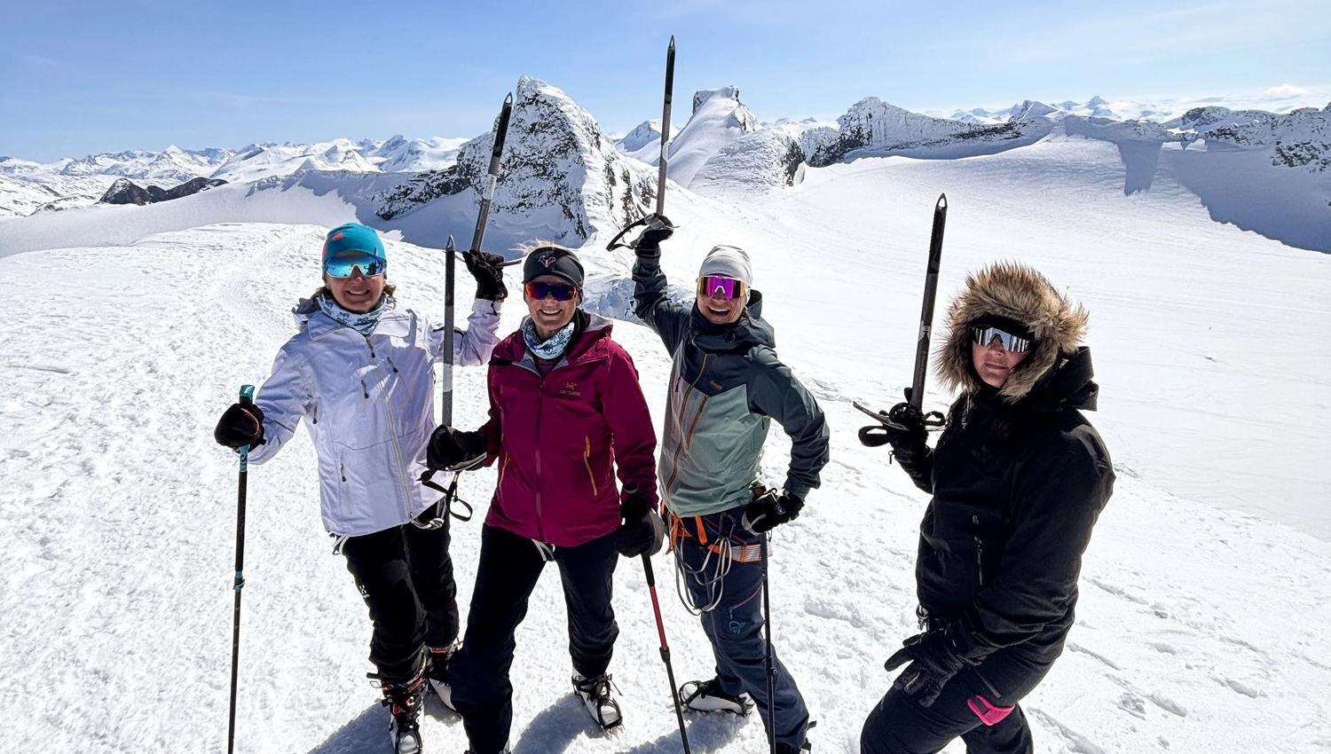 Four skiers stand on a snowy mountain ridge holding skis upright, with bright sun and rugged peaks behind them..