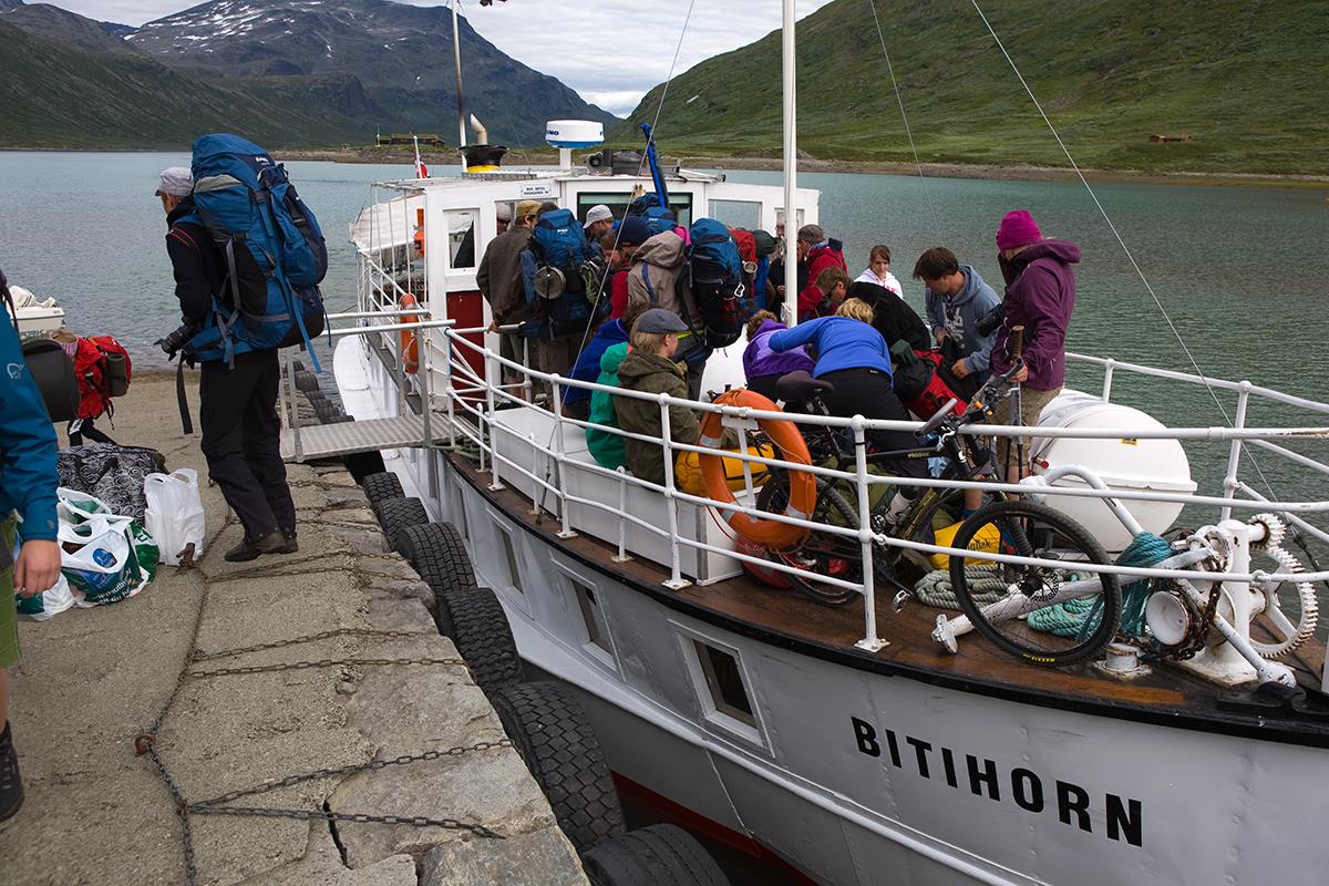 Passasjerbåt ved brygga i t fjellvann. Turfolk med ryggsekker og syklister med sykler går fra bord.
