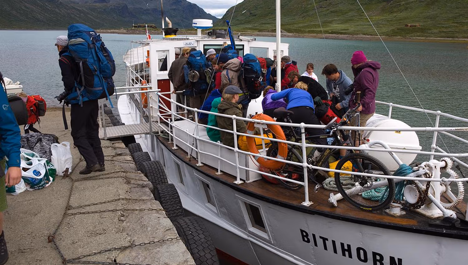 Passasjerbåt ved brygga i t fjellvann. Turfolk med ryggsekker og syklister med sykler går fra bord.