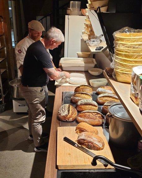 Two smiling men baking bread. Several freshly baked loaves on the counter in front of them.