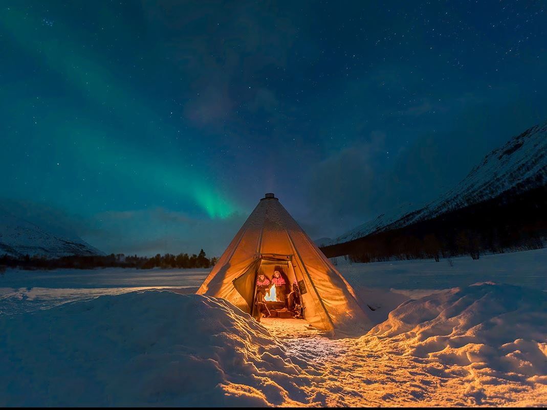 A tent with guests inside and the Northern Lights above in a winter landscape