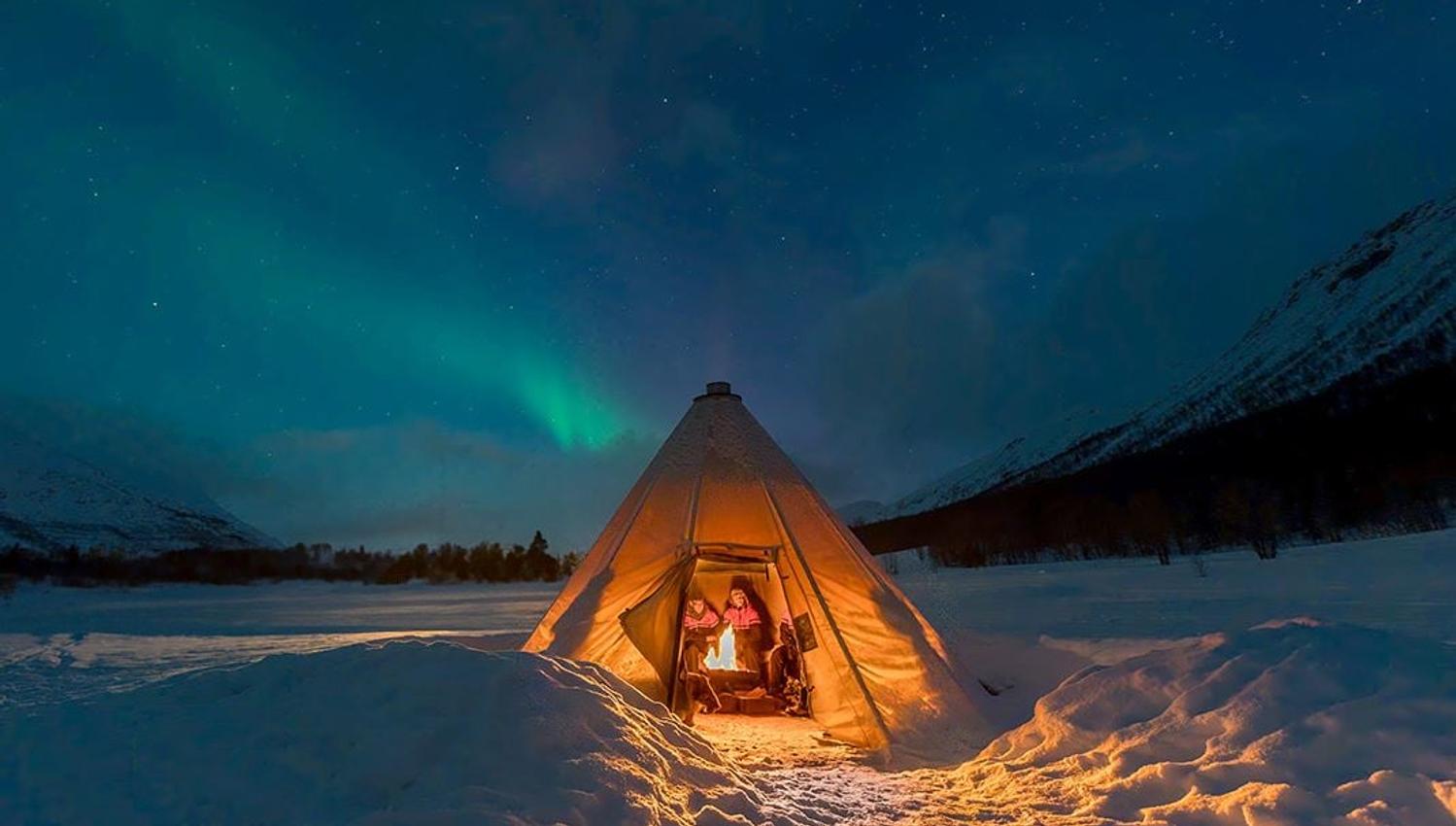 A tent with guests inside and the Northern Lights above in a winter landscape