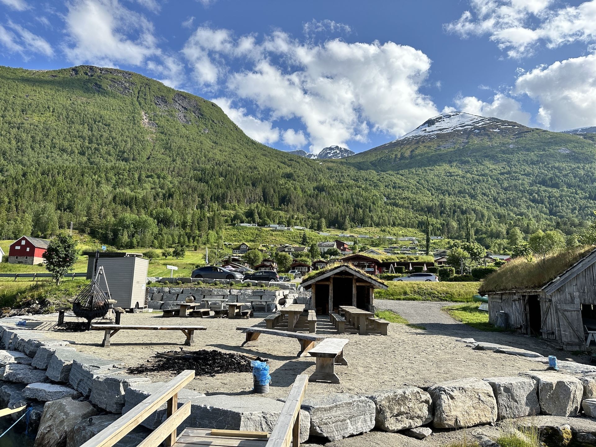 Rustic picnic benches and a shelter in a gravel yard, with green mountains and a snowy peak under clouds..