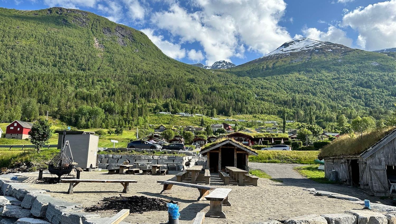 Rustic picnic benches and a shelter in a gravel yard, with green mountains and a snowy peak under clouds..