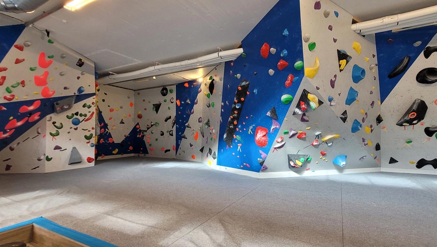 Empty indoor bouldering gym with blue and gray climbing walls covered in colorful holds and thick padded flooring..