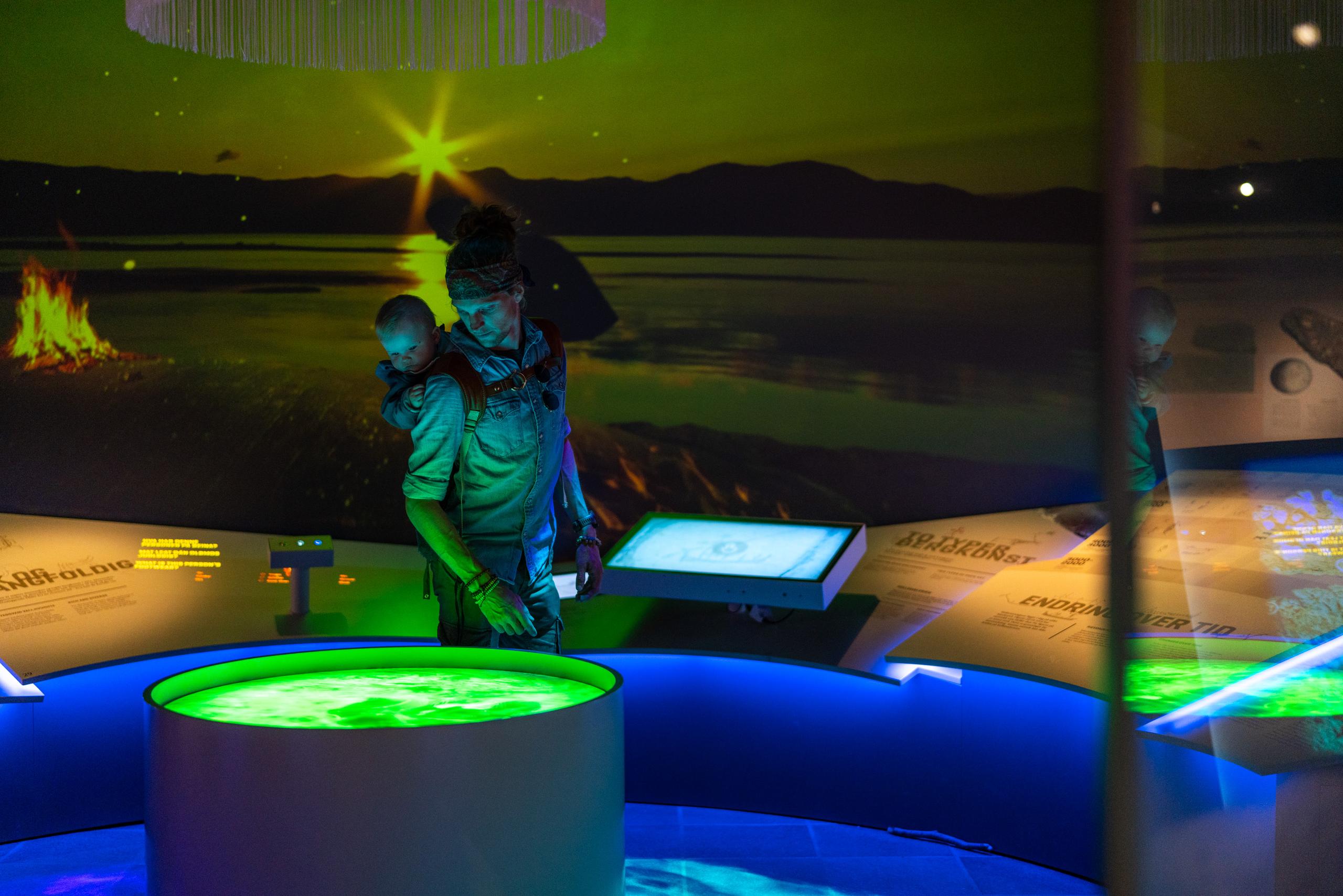 Une personne avec un enfant porte sur le dos explore une exposition interactive lumineuse.