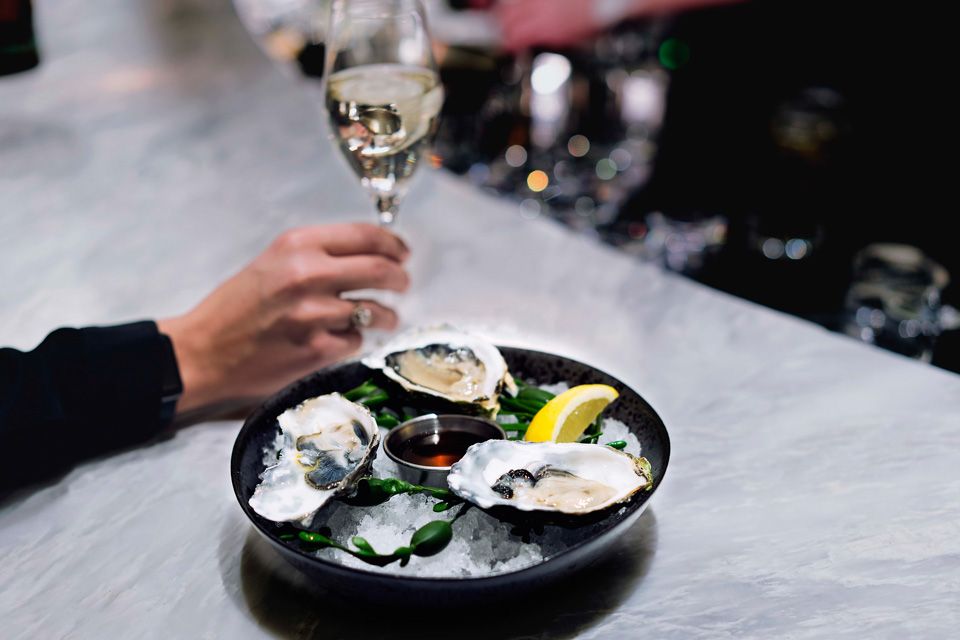 A hand holding a glass of white wine next to a plate of oysters