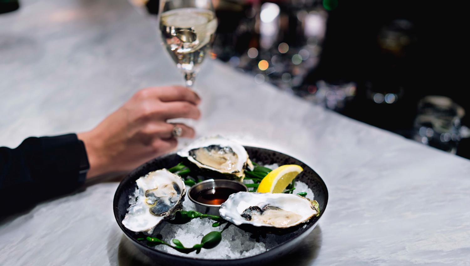 A hand holding a glass of white wine next to a plate of oysters