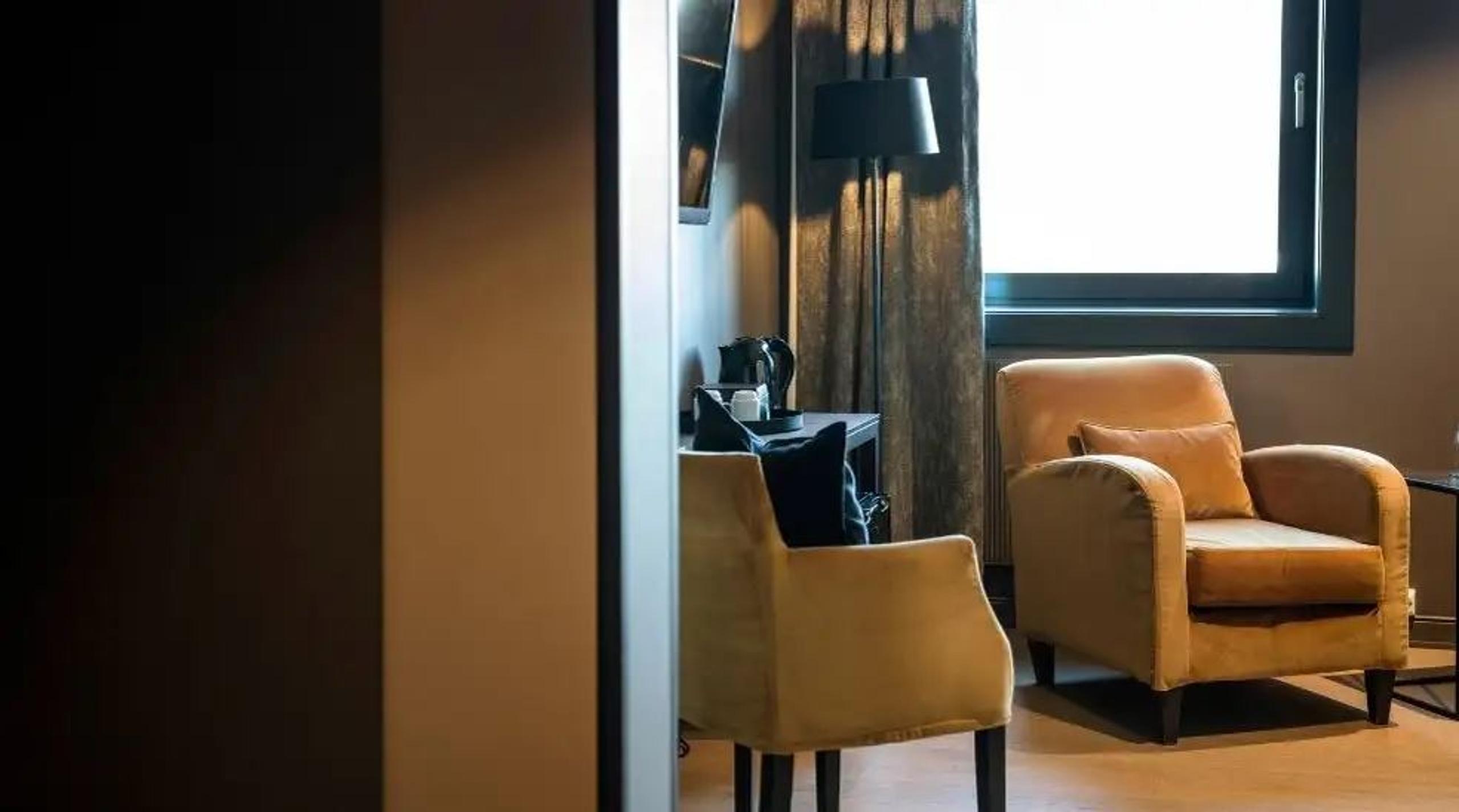 Warm-toned room with an upholstered armchair near a bright window, plus desk chair, lamp, and curtains.