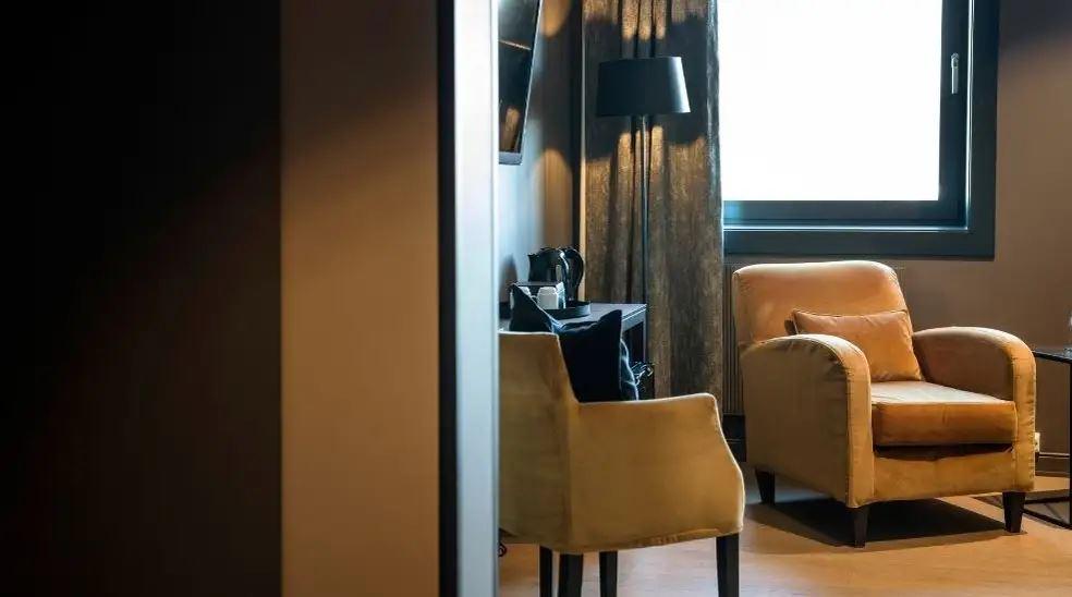 Warm-toned room with an upholstered armchair near a bright window, plus desk chair, lamp, and curtains.