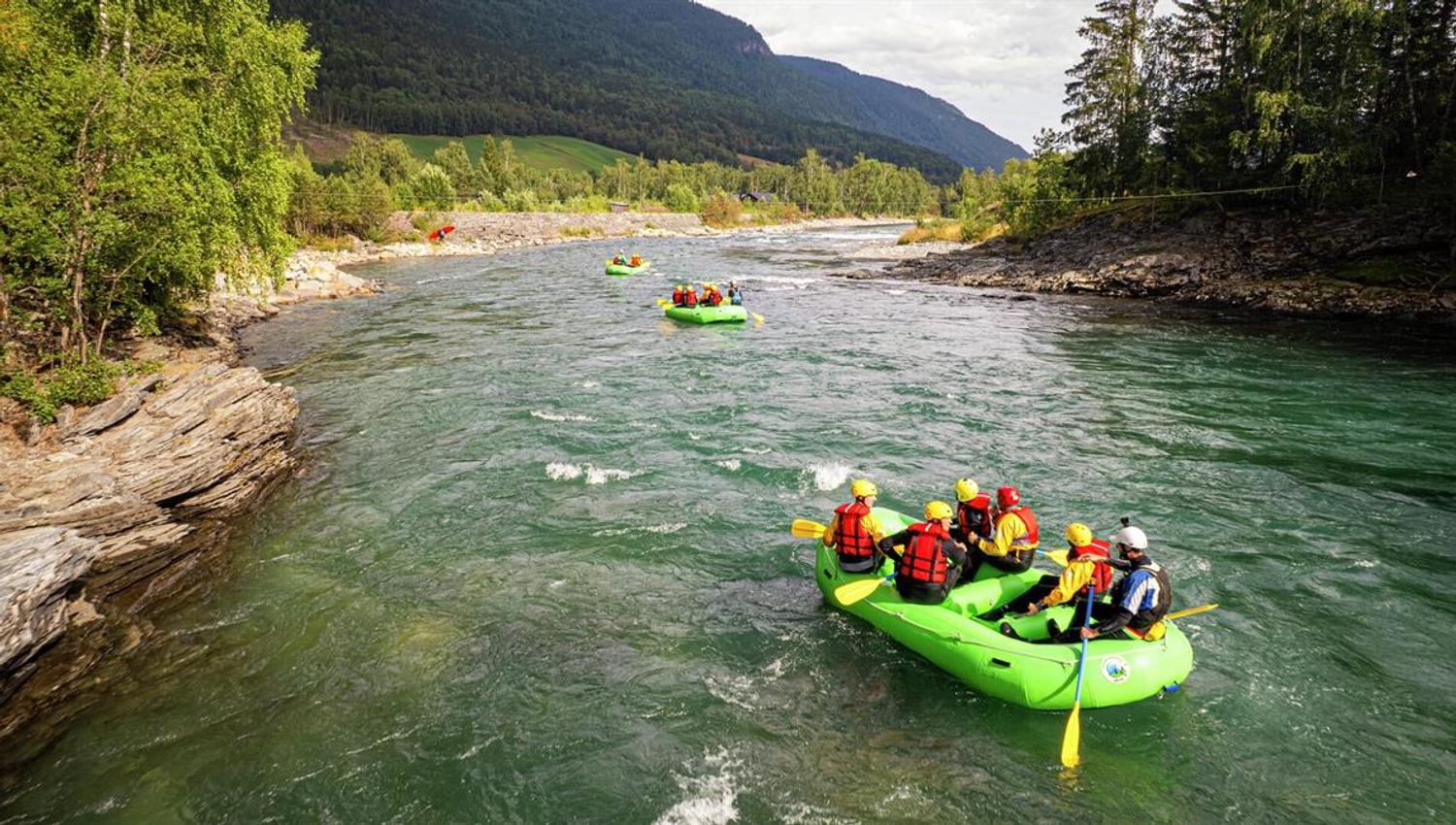 Green rafting boat with participants wearing helmets and life jackets on a calm section of the Sjoa River, surrounded by forest and mountains.