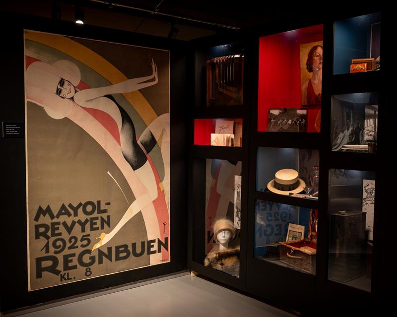 Exhibition room featuring a large poster for the 1925 Mayol revue, surrounded by display cases containing photographs, hats and other objects from the