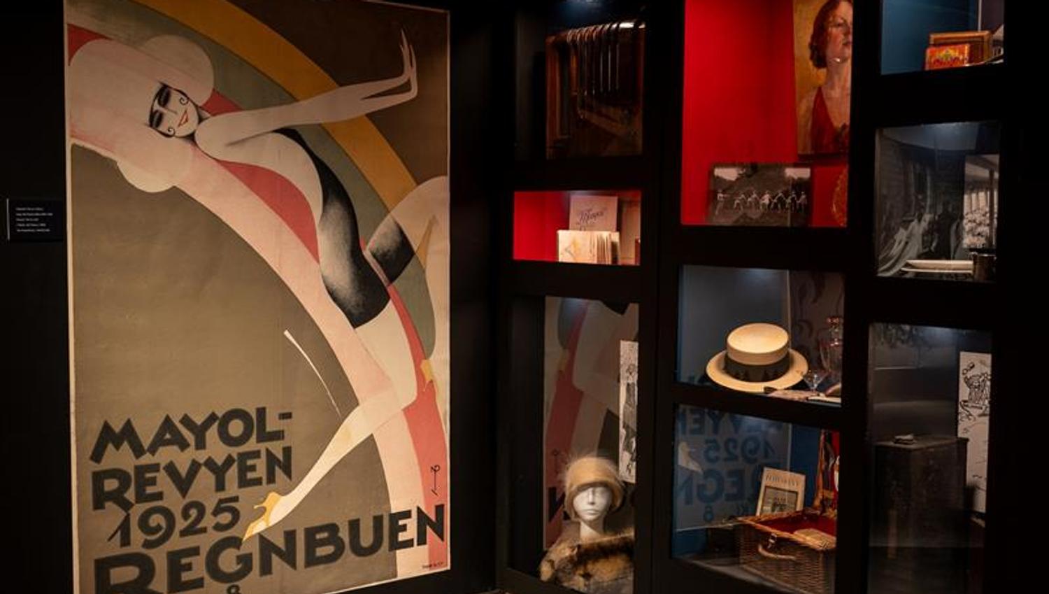 Exhibition room featuring a large poster for the 1925 Mayol revue, surrounded by display cases containing photographs, hats and other objects from the