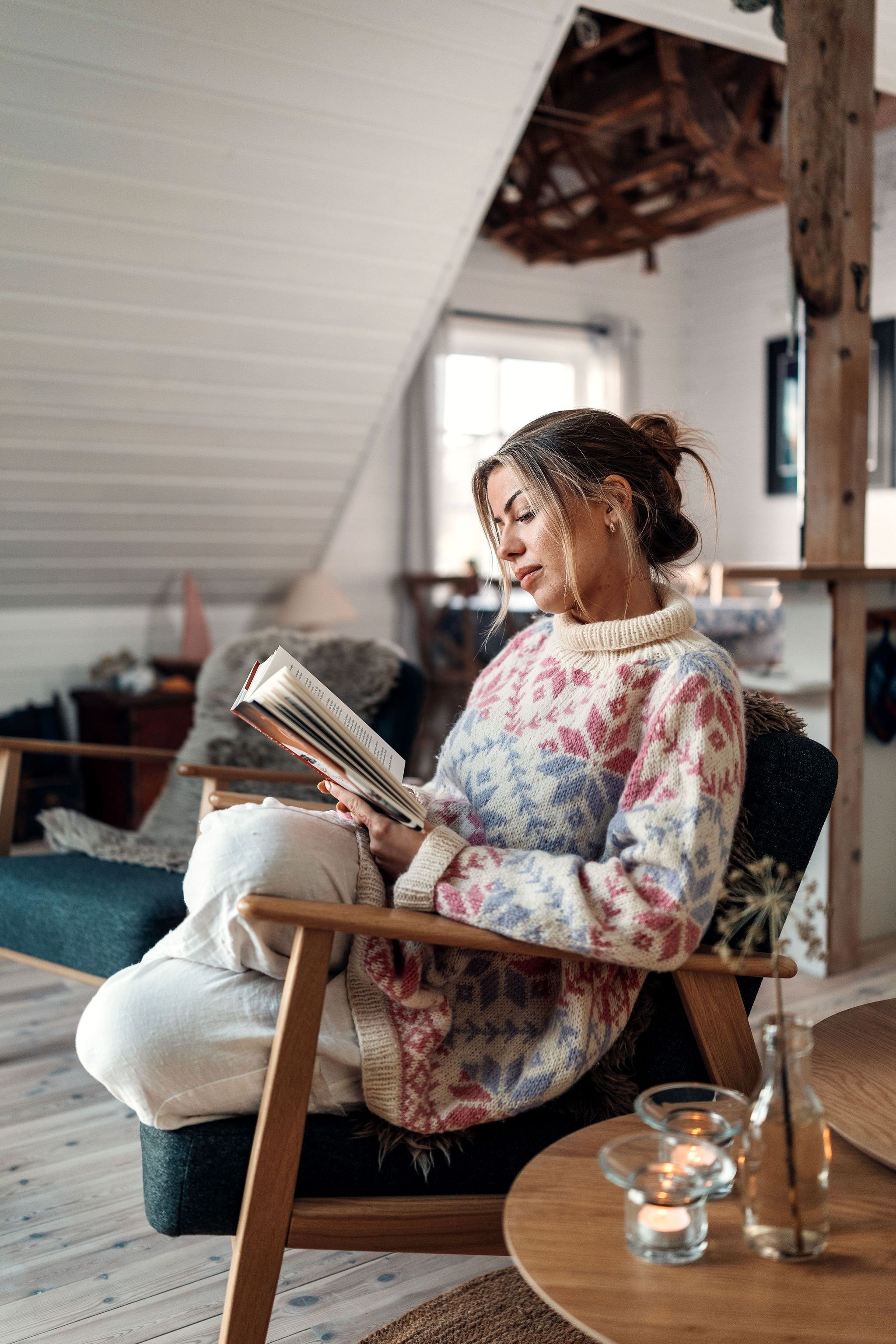 Person in a patterned sweater sits in an armchair reading a book in a loft living room with candles on a table. at the Seahouse at Hornelen Panorama in Bremanger.