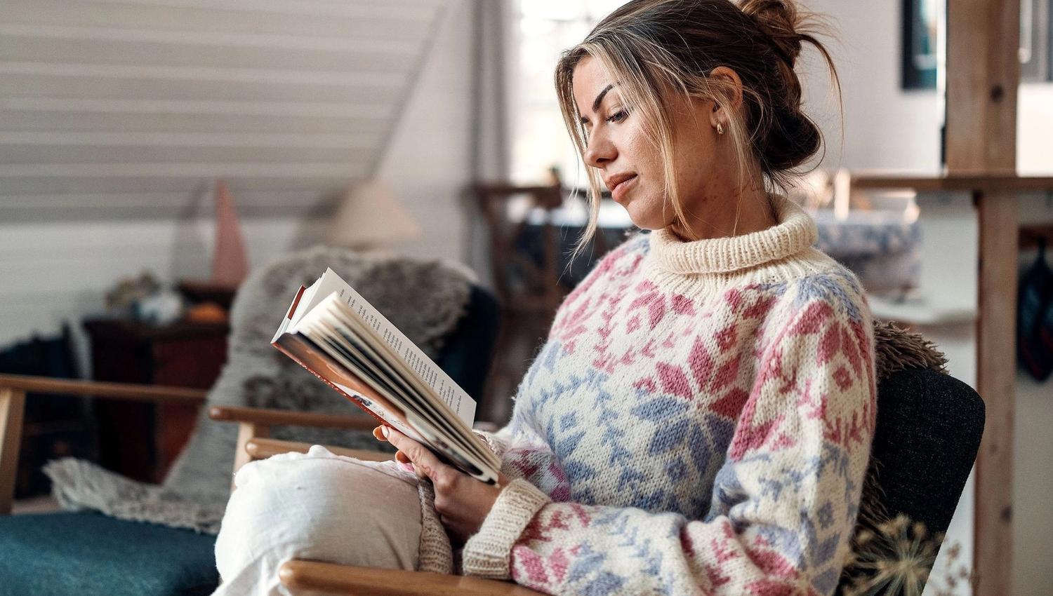 Person in a patterned sweater sits in an armchair reading a book in a loft living room with candles on a table. at the Seahouse at Hornelen Panorama in Bremanger.