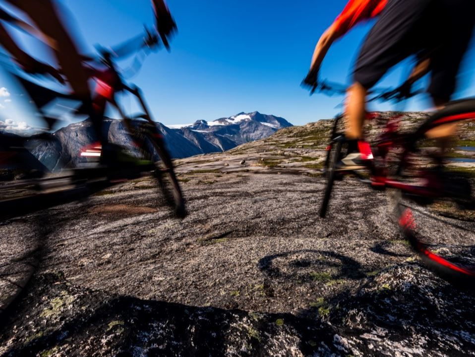 Two cyclists speeding over rocky mountain terrain, with blurred movement and mountains and blue skies in the background.