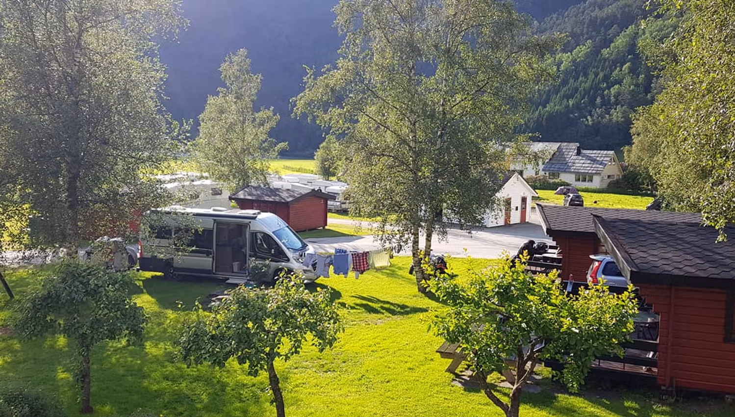 Sunny day at Folgefonn Cabins and Farm Camping in Hardanger, with red cabins, campervans, and lush green surroundings.