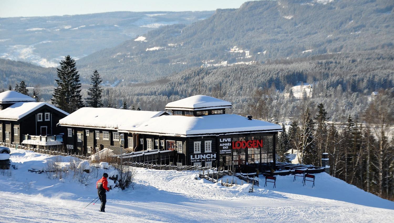 Snødekte hytter ved en alpinbakke, med en skiløper i forgrunnen og skogkledde fjell i bakgrunnen..