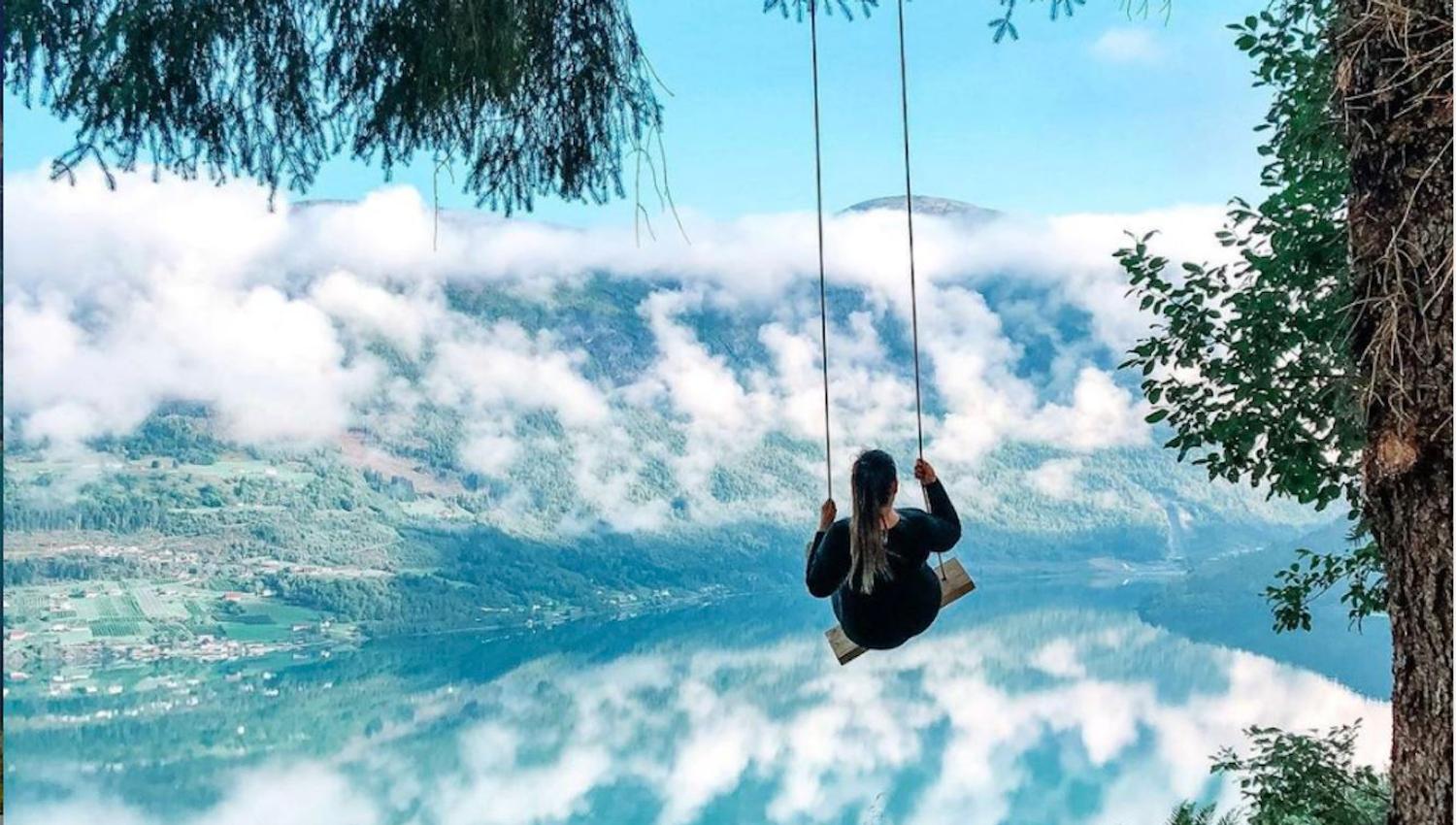 A person on a rope swing hangs from a tree above a grassy slope, overlooking a cloudy mountain lake at Skarsteinssætra i Olden.