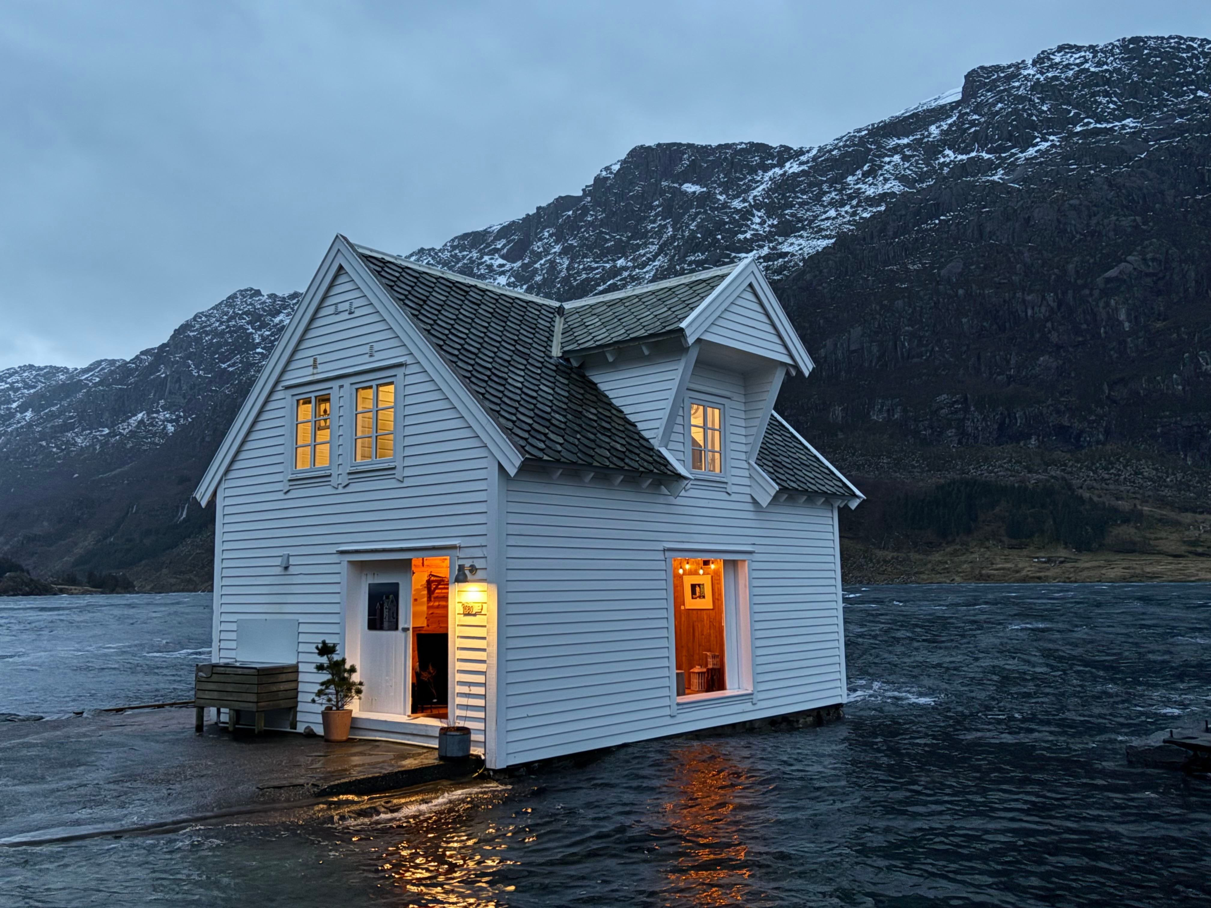 White wooden cabin with warm lit windows sits on a dock over dark water, backed by snowy mountains at dusk at the Seahouse at Hornelen Panorama in Bremanger..