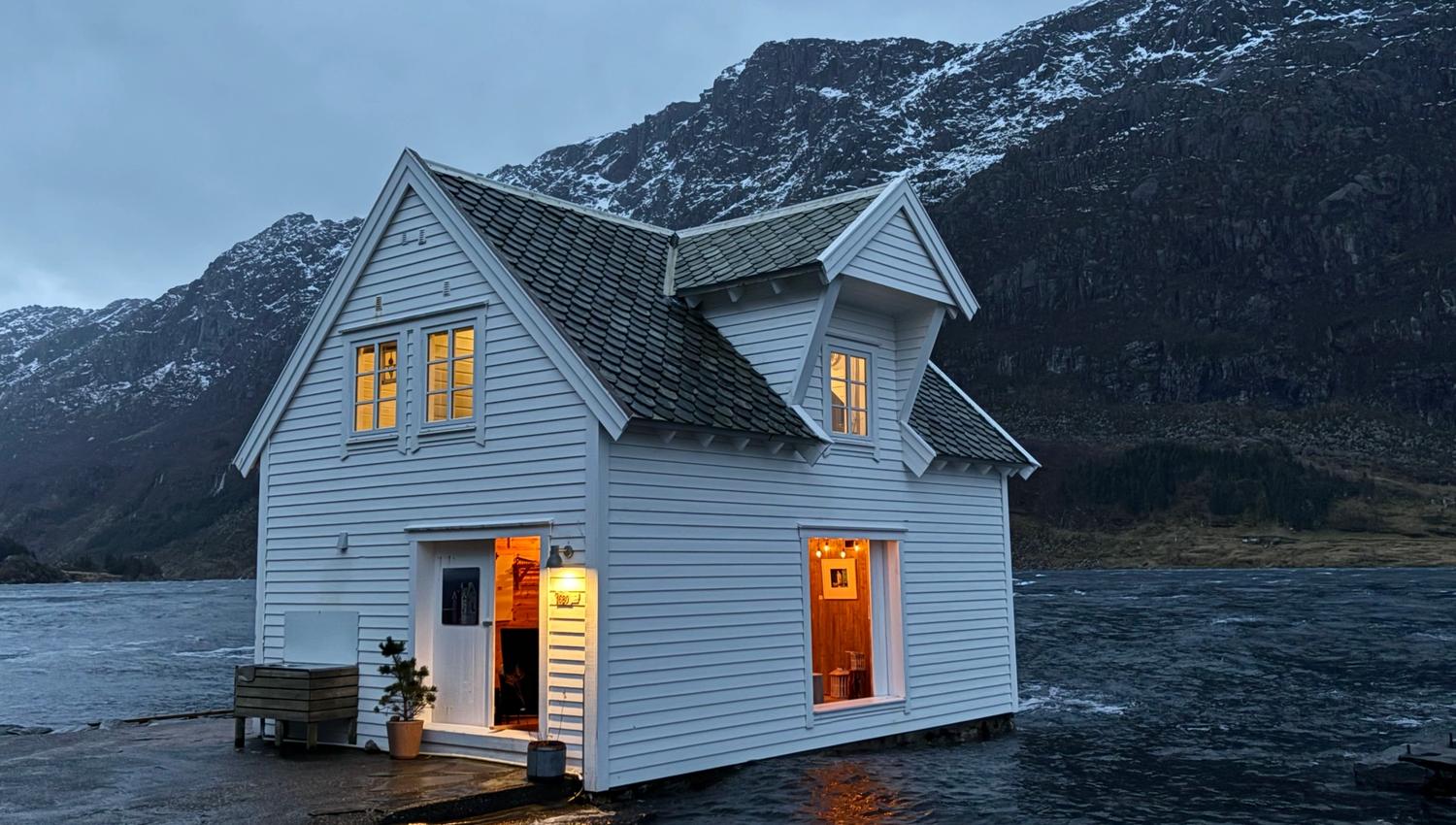 White wooden cabin with warm lit windows sits on a dock over dark water, backed by snowy mountains at dusk at the Seahouse at Hornelen Panorama in Bremanger..