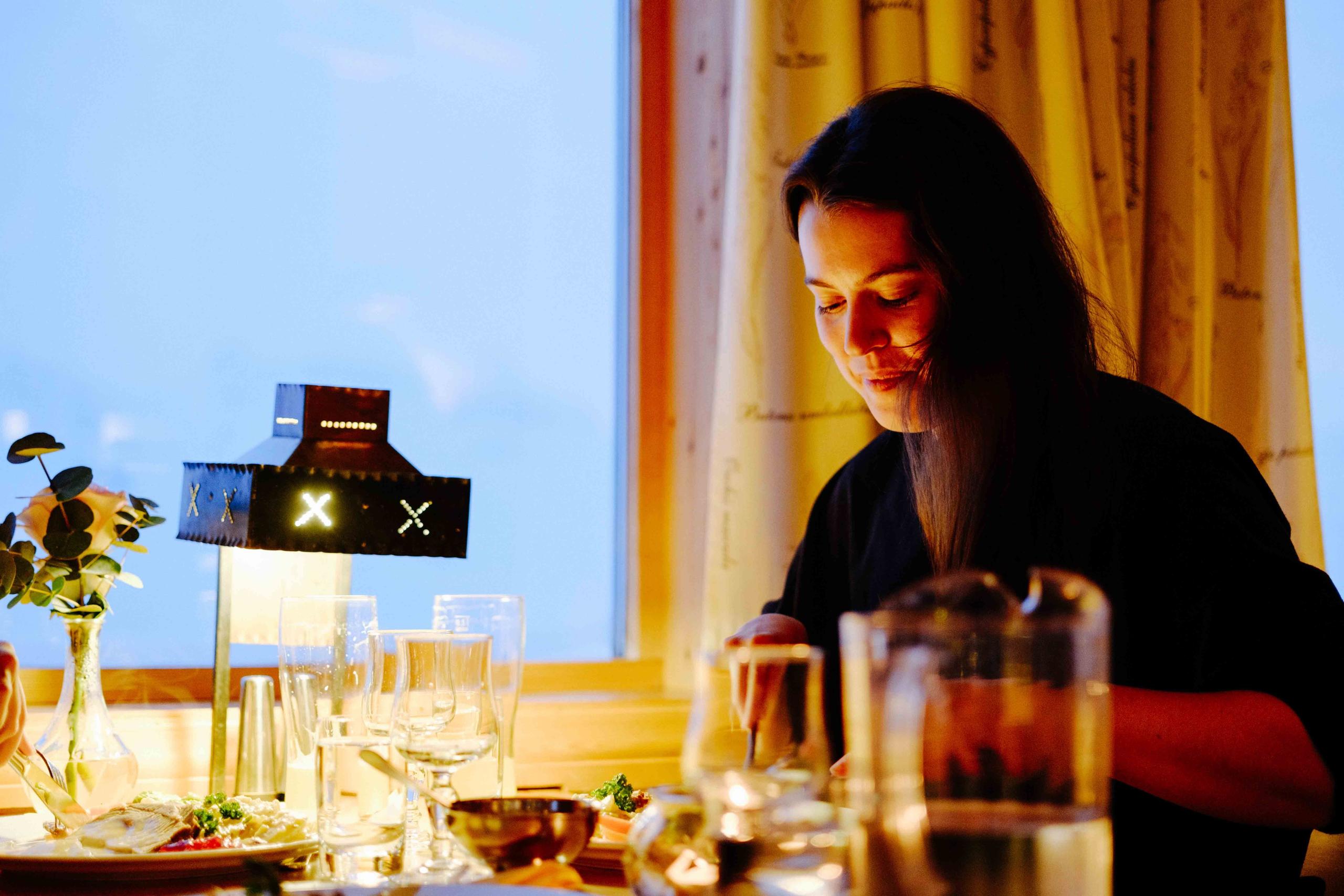 Person seated at a restaurant table with glasses and plates, lit by a small lamp beside a window..