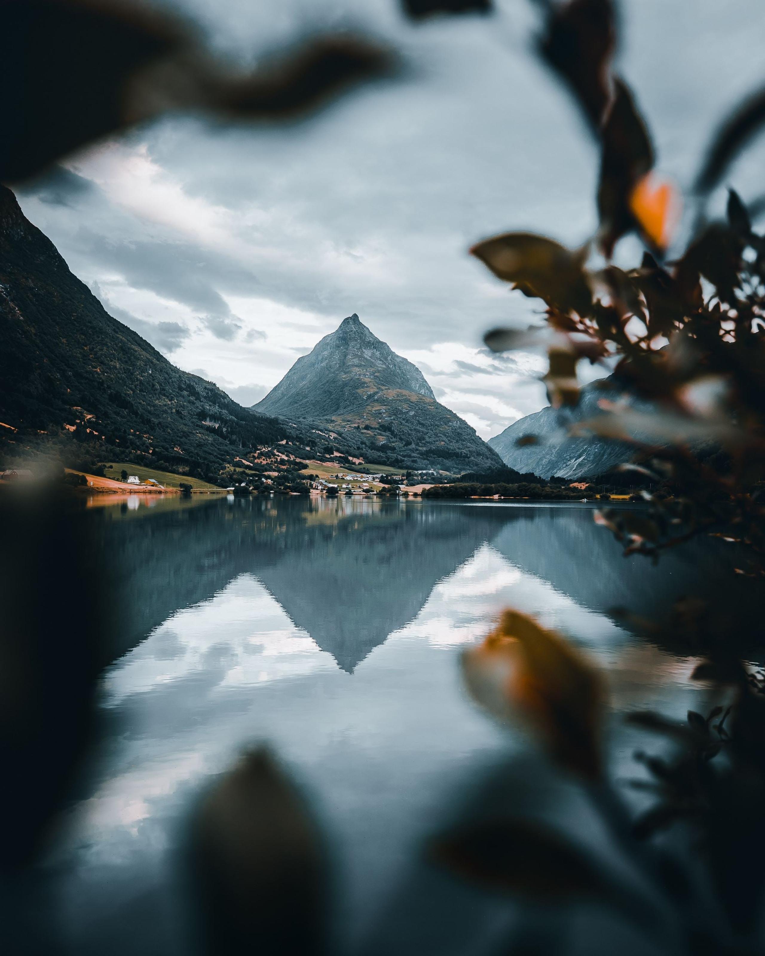 Spiss fjelltopp ved ein stille fjord med klår spegling, små bygder langs stranda og skyer på himmelen..