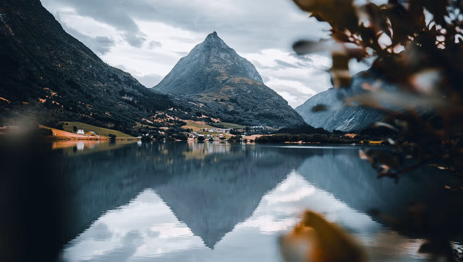 A pointed mountain rises beyond a calm lake, reflecting in the water under cloudy skies, framed by blurred leaves..